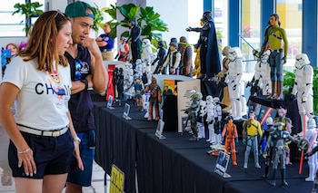 Hombre y mujer de pie, mirando una mesa cubierta con figuras de acción detalladas de Star Wars, incluyendo personajes como Darth Vader y Stormtroopers