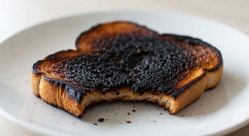 Una rebanada de pan tostado muy quemada, con un trozo mordido, descansa en un plato blanco. Migas oscuras se ven alrededor del pan.