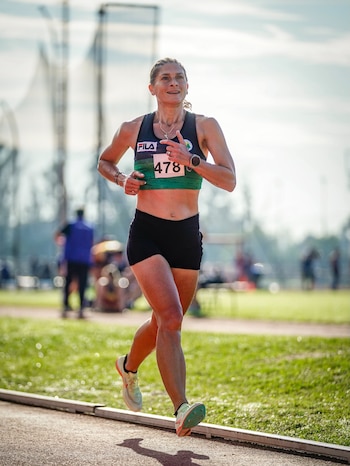 Mujer corriendo en pista de atletismo. Viste top deportivo negro y verde, pantalón corto negro, zapatillas y dorsal 478. Fondo con césped y cielo