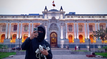 Un hombre encapuchado sostiene un teléfono celular y varias extensiones eléctricas blancas frente al edificio del Congreso de Perú con una bandera peruana.