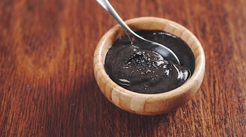 Un cuenco pequeño de madera con salsa cremosa negra y una cuchara de metal reposa sobre una superficie de madera oscura.