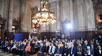 Interior de un salón con columnas, candelabro, personas sentadas y de pie, bandera argentina, cámaras de video en el fondo.