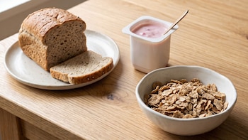 Un plato con pan Graham y dos rebanadas, un envase con yogur y una cuchara, y un cuenco con hojuelas de cereal sobre una mesa de madera