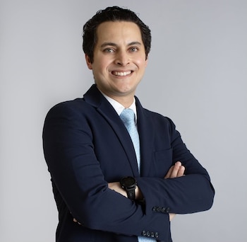 Hombre de tez clara, cabello oscuro, sonrisa, vestido con traje azul marino, camisa blanca y corbata celeste, con los brazos cruzados y un reloj negro