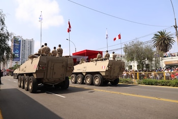Dos vehículos militares blindados de color arena con soldados, bandera de Perú, público y edificios en la Av. Brasil bajo cielo claro