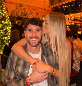 Una mujer rubia besa la mejilla de un hombre sonriente con barba, con sus brazos entrelazados; detrás, un árbol de Navidad iluminado y decorado