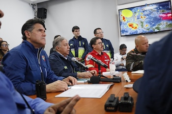 Hombres en una reunión con micrófonos y documentos sobre la mesa. Una pantalla grande al fondo muestra un mapa meteorológico con nubes y sistemas de tormentas