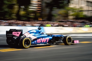 Coche de carreras azul y rosa con 'BWT' en el lateral, moviéndose a alta velocidad en una pista de asfalto. Fondo borroso con espectadores, árboles y edificios