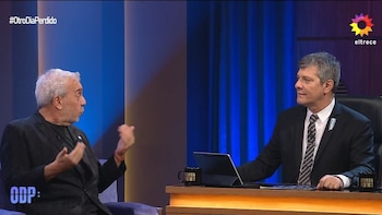 Dos hombres en un estudio de televisión. Uno de pelo blanco gesticula; el otro, de traje, está sentado frente a un escritorio con una laptop y dos tazas negras