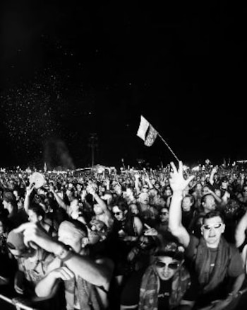 Multitud en blanco y negro con brazos levantados. Una bandera ondea. Personas usan gafas de sol. Luces difusas en el fondo contra cielo oscuro