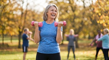 Mujer de más de 50 años con camiseta azul y mallas negras, sonriente y sosteniendo mancuernas rosas en un parque con árboles de otoño.