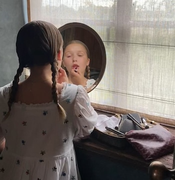 Vista posterior de una niña con trenzas y un vestido blanco bordado mirándose en un espejo redondo, aplicándose lápiz labial. Artículos de maquillaje sobre la mesa