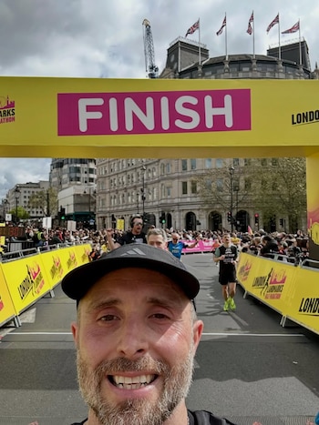 Hombre barbudo sonriendo en primer plano, se toma una selfie en la meta de la London Landmarks Half Marathon, con corredores y público al fondo