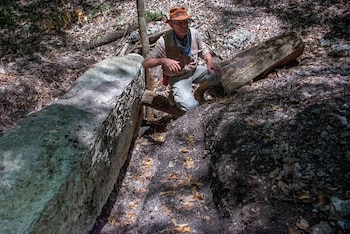 Un hombre con sombrero, pañuelo, camisa clara, pantalón y botas se agacha entre grandes rocas grises, tierra, hojas secas y ramas de árboles en un bosque