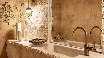 Lavabo de piedra con grifo de bronce, espejo, cuenco con cactus, dos jarrones con ramas y un aplique de luz en pared de roca.