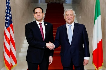 Dos hombres con traje se dan la mano frente a las banderas de Estados Unidos, Italia y la Unión Europea, con una escalera roja al fondo