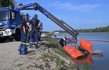 Varios rescatistas junto a un camión azul con brazo articulado en una orilla mientras una persona en el agua trabaja con flotadores naranjas