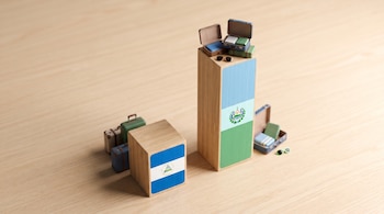 Dos bloques de madera con banderas de Nicaragua y El Salvador sobre una mesa, uno bajo con maletas cerradas, otro alto con maletas abiertas y gafas de sol.