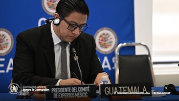 Un hombre con traje, anteojos y auriculares escribe junto a letreros con los textos Presidente del Comité de Expertos MESICIC y Guatemala