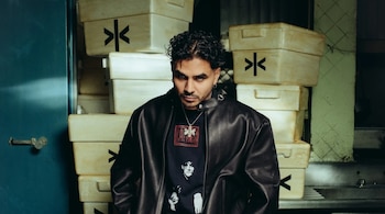 Retrato de Álvaro Díaz mirando a la cámara, con pelo rizado y barba, vistiendo chaqueta de cuero negra y camiseta gráfica, frente a cajas apiladas