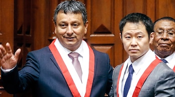 Primer plano de tres hombres: dos con trajes oscuros, camisas blancas, corbatas y bandas ceremoniales rojas y blancas; el tercero detrás con lentes. Fondo de madera.