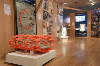 Una maqueta tubular de color naranja en un pedestal blanco, rodeada de paneles con fotografías arquitectónicas y suelo de madera