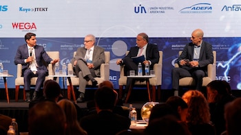 Cuatro hombres en traje sentados en un escenario, participando en un panel de discusión con micrófonos y botellas de agua. Una pantalla con logos al fondo
