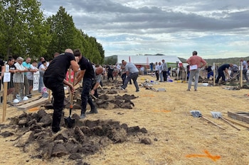 Varios hombres excavan la tierra con palas en un terreno al aire libre mientras personas observan detrás de una valla metálica