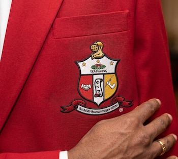 Blazer rojo con escudo bordado en el bolsillo: yelmo, apretón de manos, pergamino con letras, antorcha, estrellas y texto griego. Una mano con anillo de oro