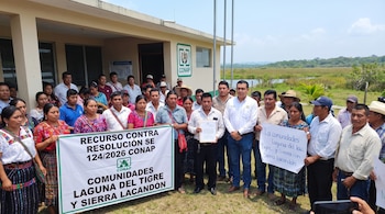 Un grupo de personas sostiene dos letreros y un documento frente a un edificio con el logo de CONAP, con un humedal y vegetación al fondo.