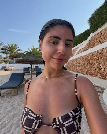 Una mujer joven con cabello mojado y bikini a rayas se toma una selfie en la playa. Al fondo, palmeras y tumbonas de playa