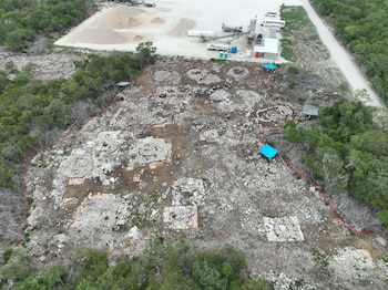 Vista aérea de un terreno delimitado con cimentaciones de piedra, personas con chalecos reflectantes, carpas azules y vegetación