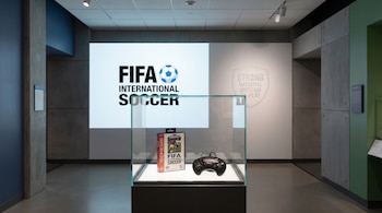 Sala de museo con una vitrina central que contiene el videojuego FIFA International Soccer para Sega Genesis y un joystick. Al fondo, el logo del juego y del Strong National Museum of Play.