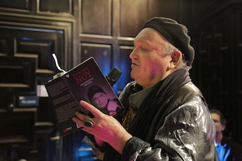 Primer plano de Performance M, un hombre con boina, bufanda y una chaqueta brillante, leyendo un libro con el título "Fernando Noy" en un micrófono