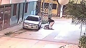 Una persona agachada auxilia a otra tendida en la vía pública junto a un automóvil aparcado frente a varias viviendas
