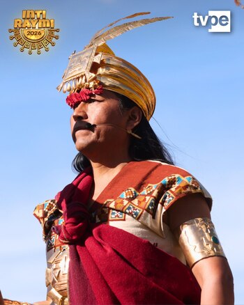 Primer plano de un hombre ataviado con vestimenta tradicional incaica, tocado dorado con plumas, túnica, fajín rojo, brazalete dorado y micrófono. Cielo azul de fondo