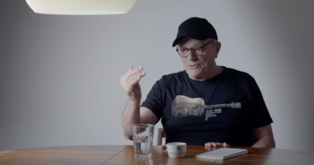 Hombre con gorra negra y gafas en mesa de madera. Viste camiseta negra con gráfico de guitarra. Junto a él, un vaso de agua y una taza
