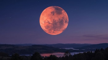 Un planeta grande y rojizo con superficie irregular en un cielo nocturno oscuro con estrellas sobre un paisaje de colinas, valles, árboles y lagos.