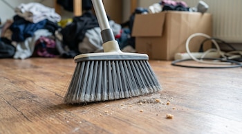 Plano detalle de una escoba gris y blanca barriendo un piso de madera laminada con polvo y residuos. Al fondo, se observan objetos y ropa desordenados.