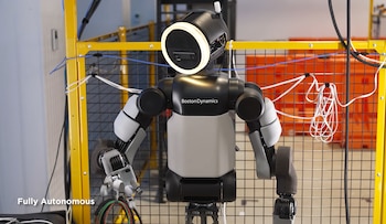 Un robot humanoide de color gris y negro con una luz circular en la cabeza está frente a una cerca metálica