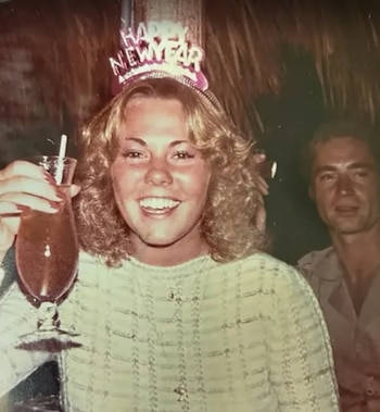 Una mujer rubia sonríe y sostiene un trago, lleva un gorro con la inscripción Happy New Year. Un hombre mira al fondo borroso