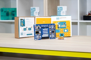 Grupo de estudiantes y adultos posan en un aula moderna con estantes llenos de kits de robótica. Sobre una mesa, un modelo de grúa y componentes tecnológicos