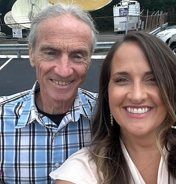 Primer plano de un hombre mayor canoso y una mujer joven con cabello oscuro sonriendo a la cámara; al fondo, antenas parabólicas y una furgoneta de noticias