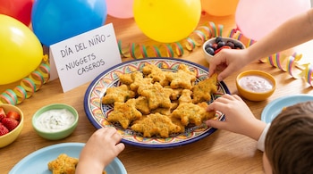 Mesa de madera con un plato de nuggets caseros con formas de estrella y dinosaurio, cuencos con fresas, arándanos y salsas. Globos y serpentinas coloridas. Manos de niños.