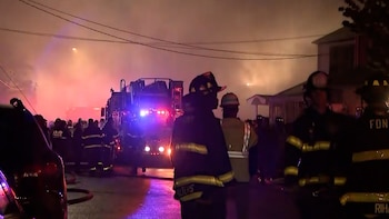 Vista nocturna de bomberos y camiones de bomberos con luces intermitentes, rodeados de una densa humareda anaranjada y rojiza en una calle urbana