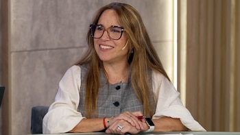 Una mujer de cabello castaño claro y gafas sonríe mientras está sentada en una mesa, vestida con una blusa blanca y un chaleco a cuadros gris