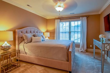 Un dormitorio con cama doble, cabecero tapizado, mesas de noche, lámparas, ventana con persianas, cortinas blancas, televisor y ventilador de techo