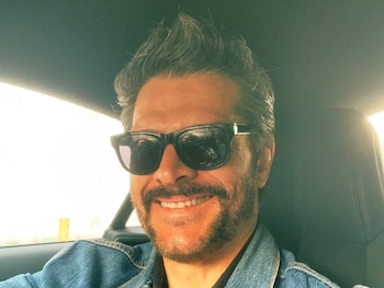 Un hombre con cabello oscuro y barba, lleva gafas de sol, una camisa polo negra y una chaqueta de denim azul, sentado en el interior de un coche