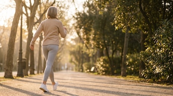 Una persona de espaldas con chaqueta beige, leggings grises y zapatillas blancas camina por un sendero arbolado. Luz del sol ilumina los árboles y el camino.