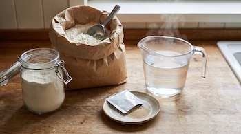 Una bolsa de papel con harina, un frasco de vidrio, una jarra de agua humeante y un plato con levadura sobre madera.